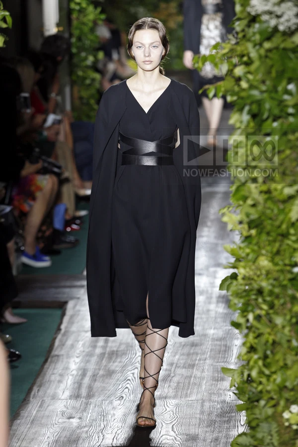 Model walking in a black midi dress with a cape overlay, a wide leather waist belt, and lace-up gladiator sandals.