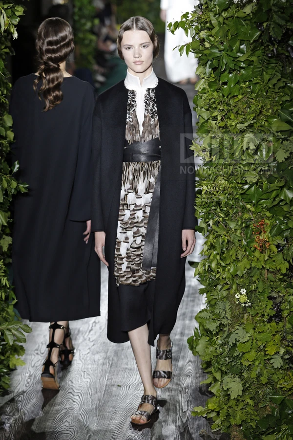 Model wearing a black coat over a feather-print dress with a wide leather waist belt, walking on a runway.
