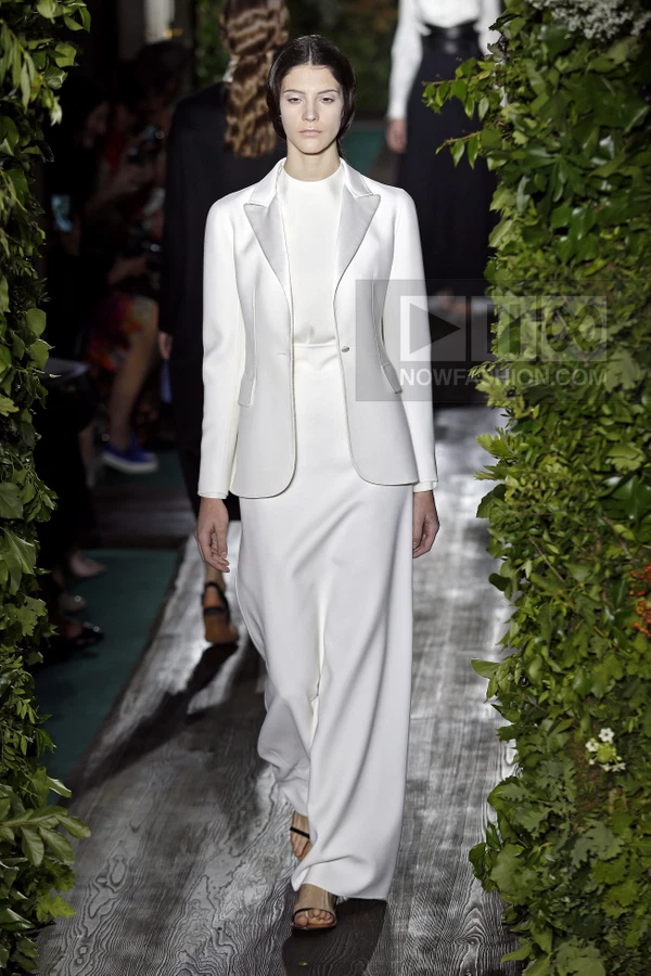 A model walks a runway wearing a white blazer over a matching floor-length dress, framed by lush green foliage.
