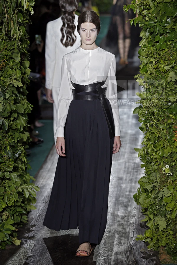Model walking on a runway wearing a long black pleated skirt, a white long-sleeve shirt, and a wide black leather belt.