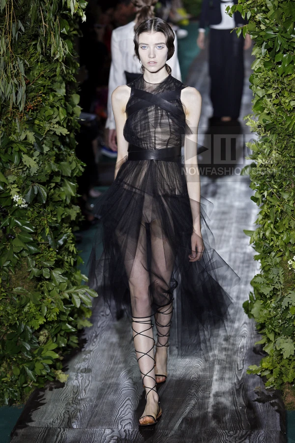Model walking on a runway wearing a black tulle dress with leather straps and lace-up sandals.