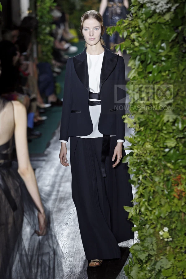 Model wearing a black blazer over a white top and long black pleated skirt on a runway with greenery.