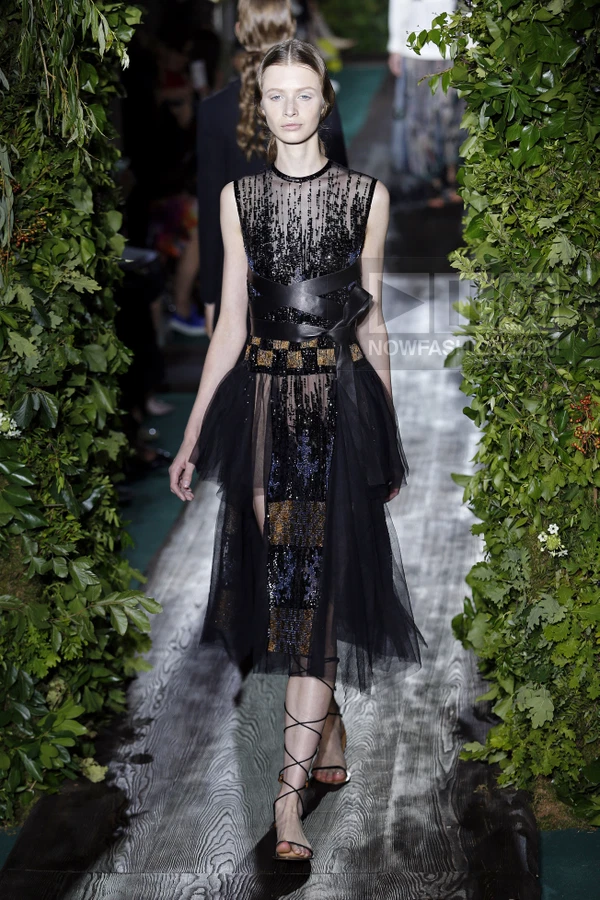 Model walking in a black sleeveless midi dress with sequin embellishments, a leather waist tie, and tulle skirt overlay.