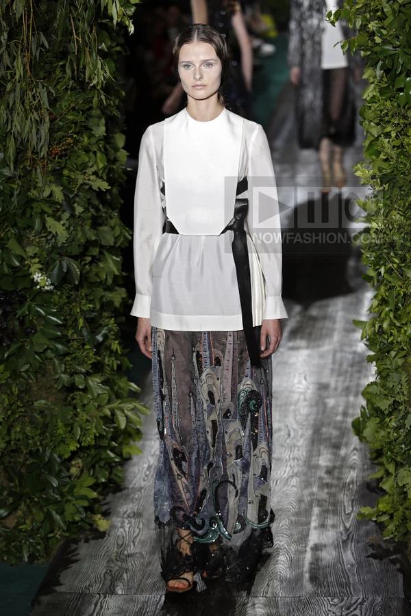 Model on runway wearing a white long-sleeve top with a black leather waist tie and a long, embroidered gray skirt.