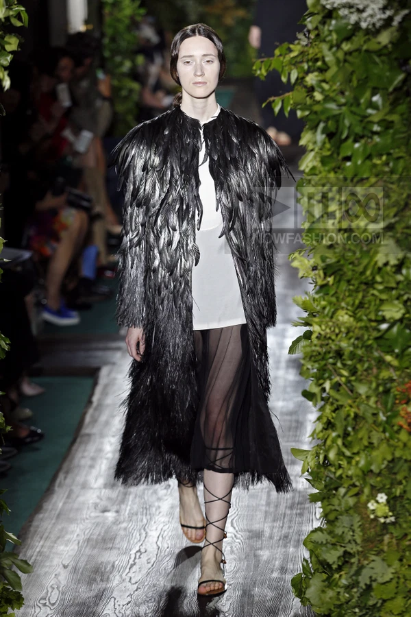 Model on runway wearing a black feathered coat over a white top and sheer black pleated skirt with lace-up sandals.