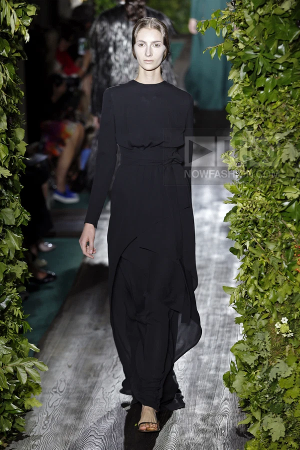 A model walks a runway wearing a long-sleeved black floor-length dress with a tied waist and layered skirt detail.
