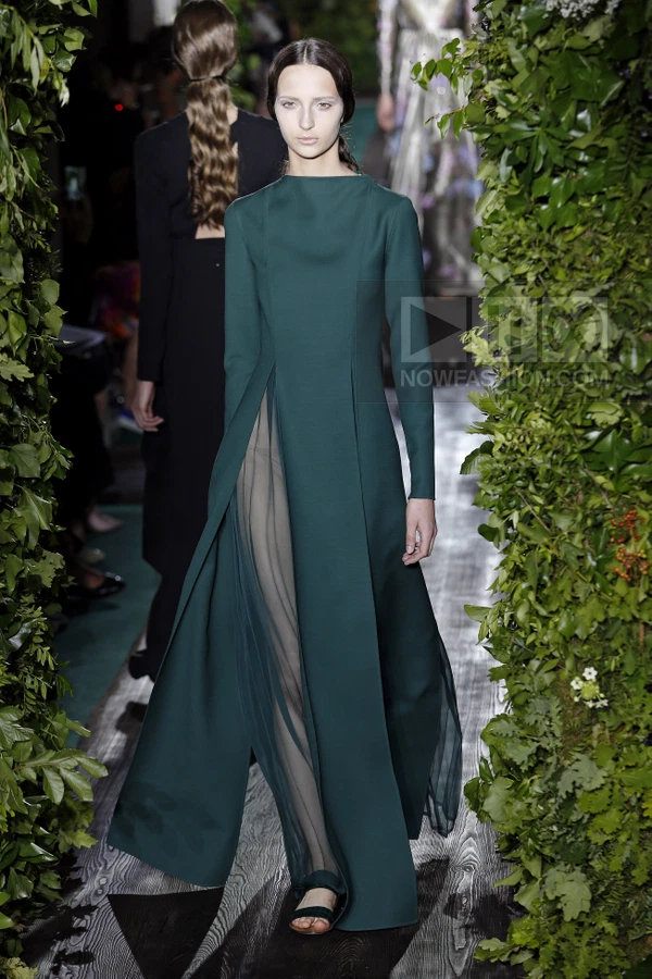 A model walks down a runway wearing a floor-length, long-sleeved dark green gown with a sheer panel at the front.