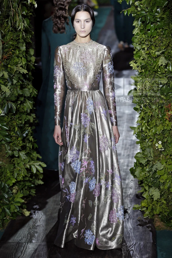 A model walks the runway in a floor-length metallic silver gown with purple and blue floral prints and lace detailing.