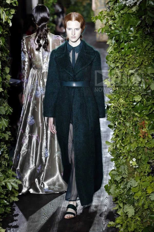 Model wearing a dark green mohair coat over a sheer dress, walking on a runway flanked by greenery.