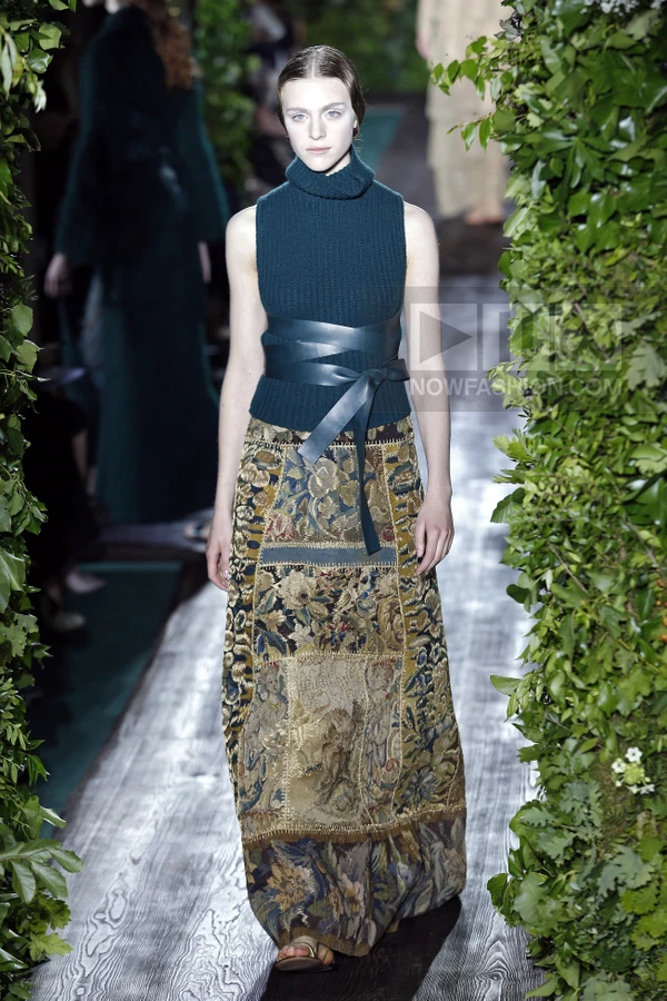 A person wearing a dark green sleeveless turtleneck sweater, a wide leather wrap belt, and a long tapestry-print skirt.
