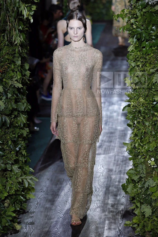 Model walking on a runway wearing a long-sleeved, tiered, sheer lace gown in a light beige color.