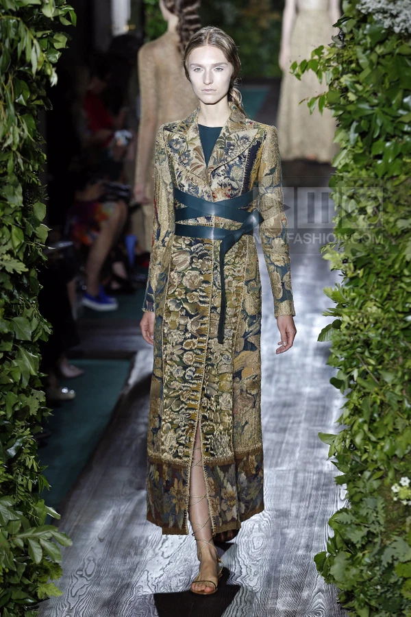 Model walking in a long, floral-patterned brocade coat with a dark green wrap belt over a dark green dress.