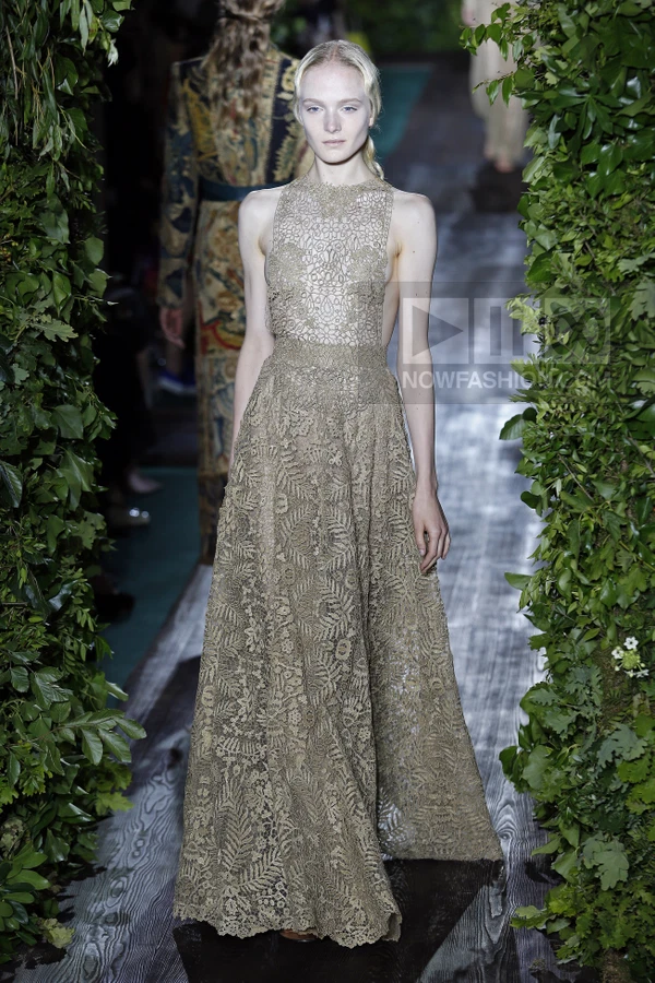 Model wearing a floor-length beige lace gown with a high neckline and open back, walking on a runway with greenery.