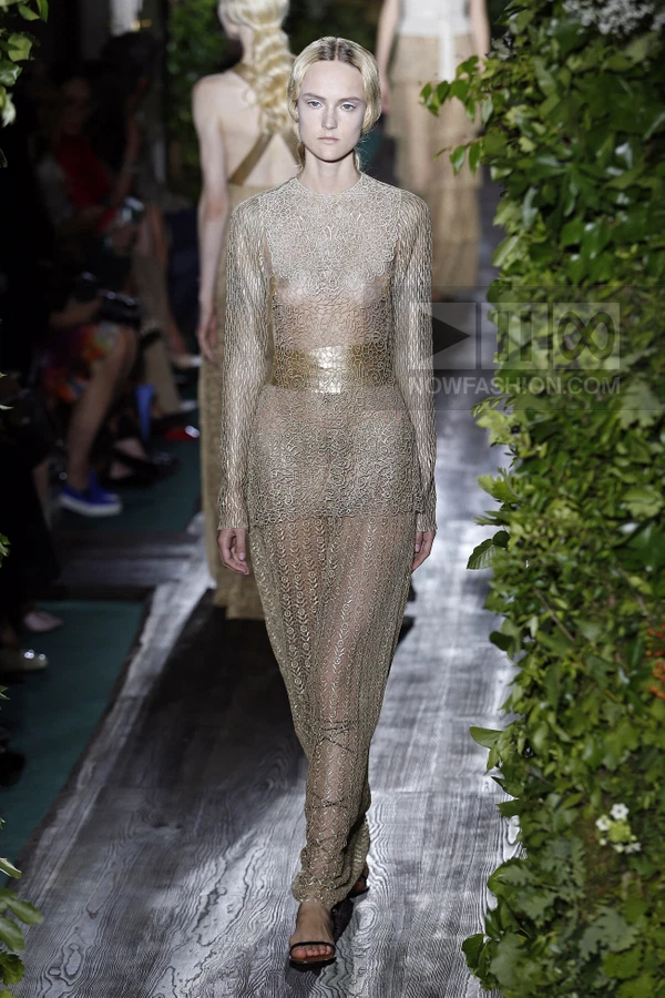 A model walks the runway in a floor-length, long-sleeved gold lace gown with a metallic waist belt.