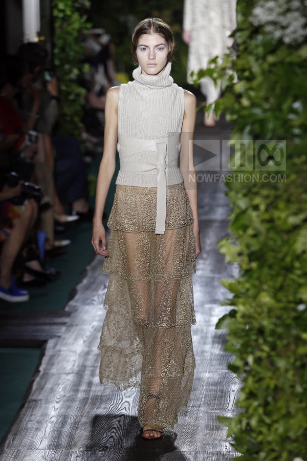 Model walking in a sleeveless ribbed knit turtleneck top and a tiered lace maxi skirt with a wide leather waist belt.