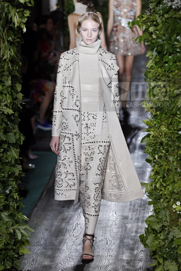 Model wearing a beige coat and matching skirt with intricate metal stud embroidery and a ribbed turtleneck sweater.