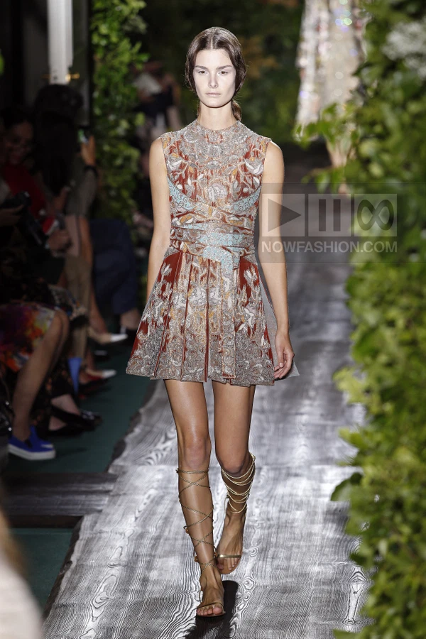 Model walking in a sleeveless mini dress with intricate rust, brown, and light blue embroidery, paired with lace-up sandals.