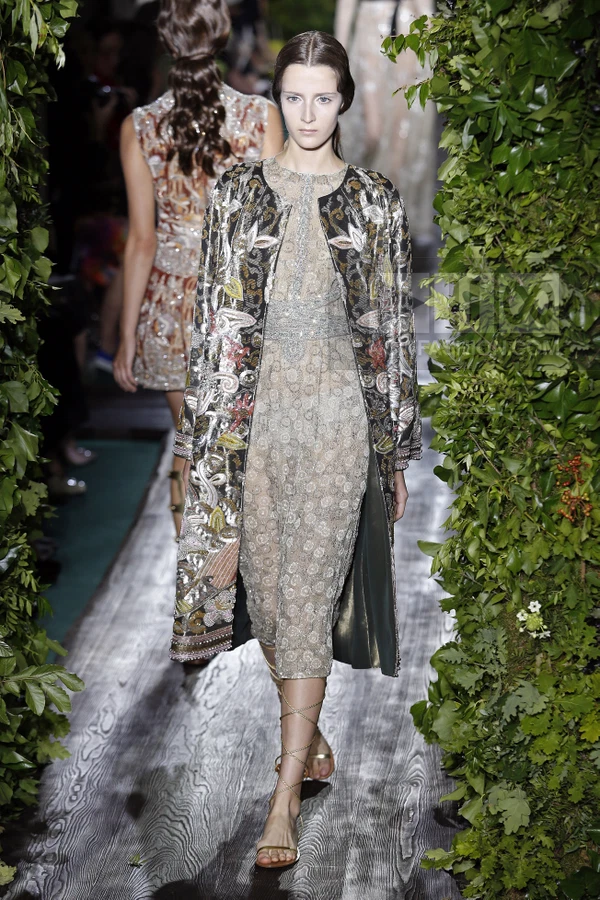 A model walks a runway wearing a long, embroidered coat over a sheer, patterned midi dress with lace-up sandals.