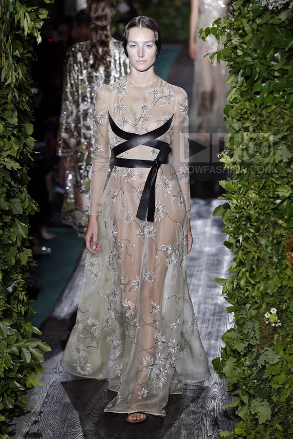 A model walks a runway in a sheer, floor-length gown with floral embroidery and black leather straps at the bodice.