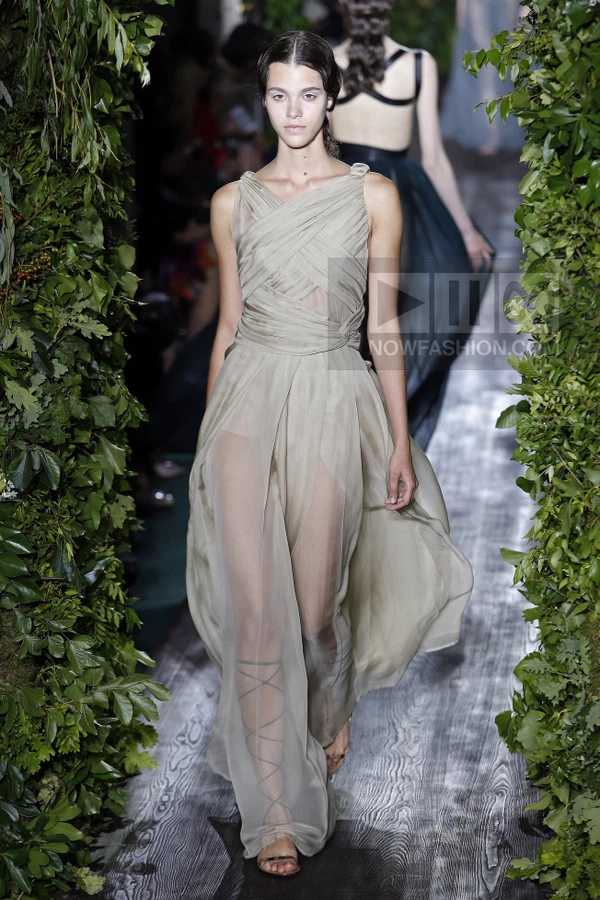 Model walking in a floor-length beige chiffon gown with a criss-cross bodice and sheer skirt on a runway.