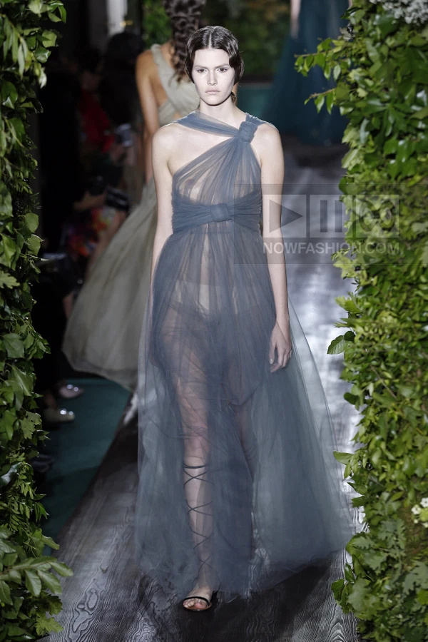 Model walking in a floor-length, sheer gray tulle gown with a knotted one-shoulder neckline on a runway.