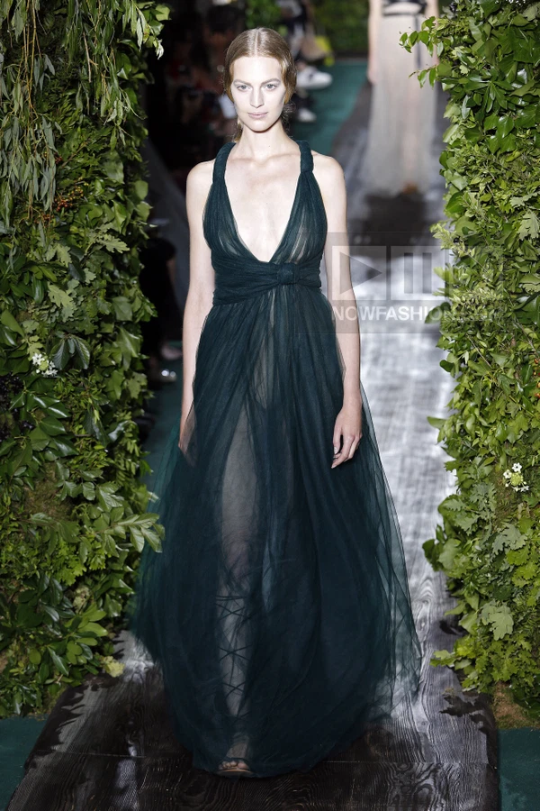 A model walks a runway in a dark green floor-length tulle gown with a plunging neckline and knotted waist detail.