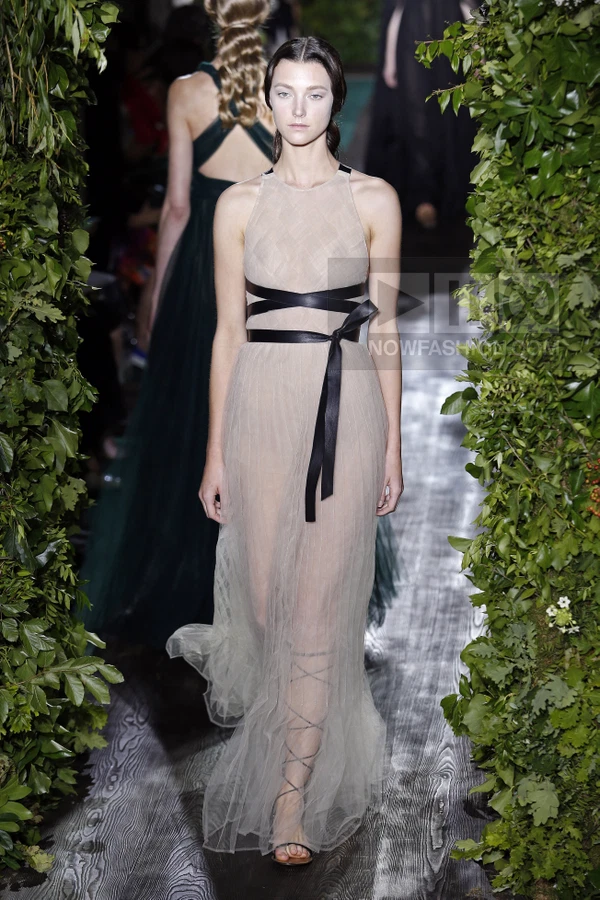 Model wearing a floor-length sheer beige tulle gown with a black leather waist tie on a runway flanked by greenery.