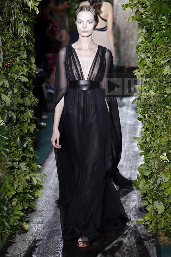 A model walks a runway in a floor-length black sheer chiffon gown with a wide leather waist belt.