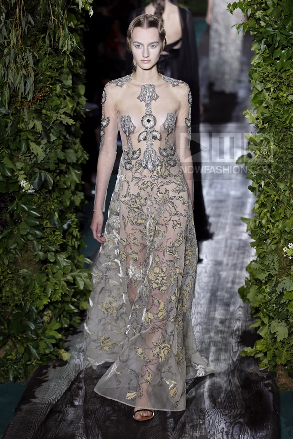 A model walks a runway in a sheer floor-length gown with silver and gold floral embroidery and long sleeves.
