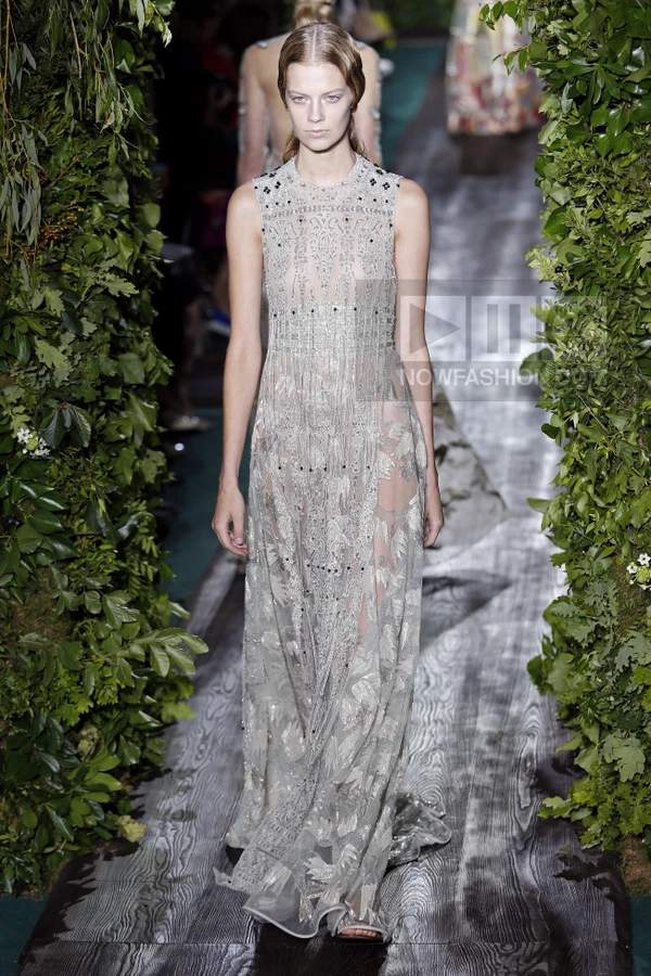 A model walks a runway in a floor-length, sleeveless, sheer silver gown with intricate embroidery and beadwork.