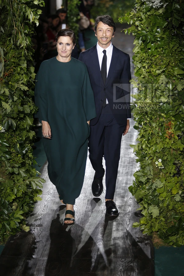 Two individuals walking down a runway flanked by lush green foliage, wearing dark green dress and navy suit.