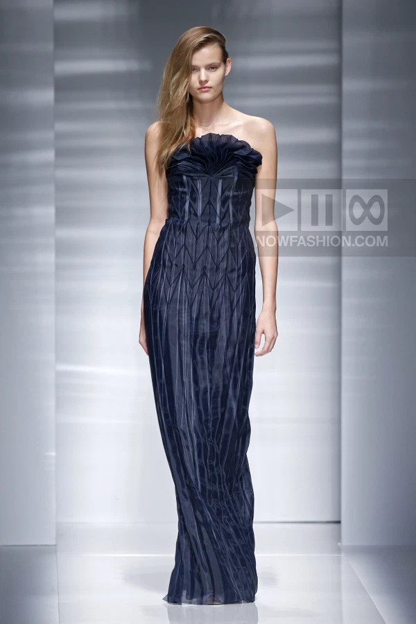 A model wearing a navy blue strapless floor-length gown with vertical pleated details on a runway.