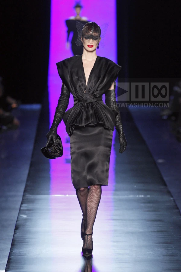 Model on runway wearing black satin dress, opera gloves, and butterfly-shaped headpiece with dark mesh veil.