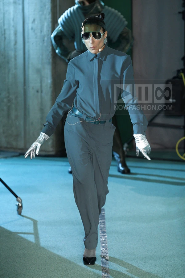 A person wearing a blue button-up shirt and matching trousers with a thin belt, silver gloves, and dark sunglasses.