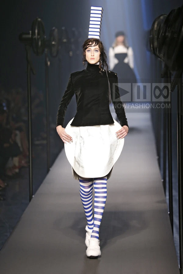Model on runway wearing black velvet jacket, white structured skirt, blue and white striped tights, and tall hat.