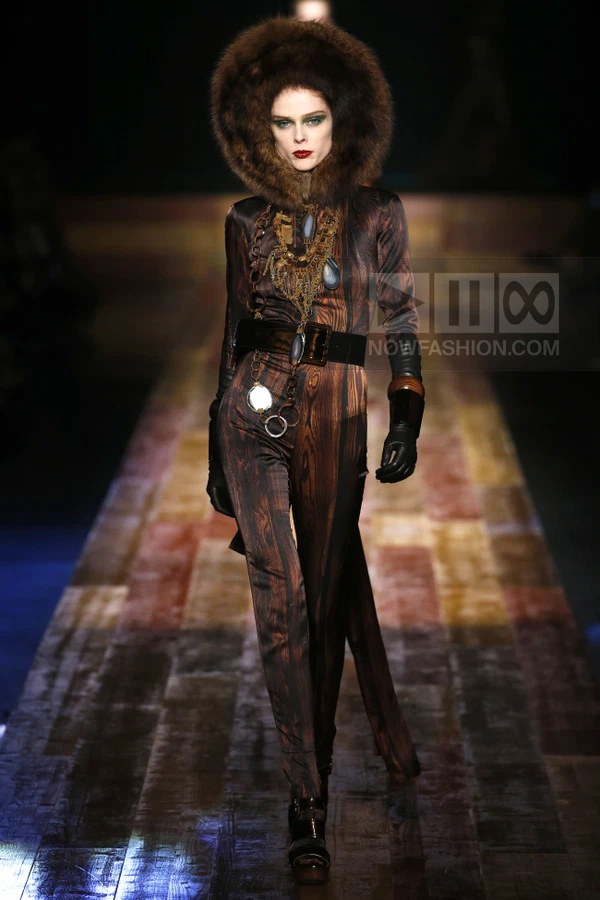 Model wearing a brown wood-grain print jumpsuit with a large fur hood, wide belt, and layered statement necklace.