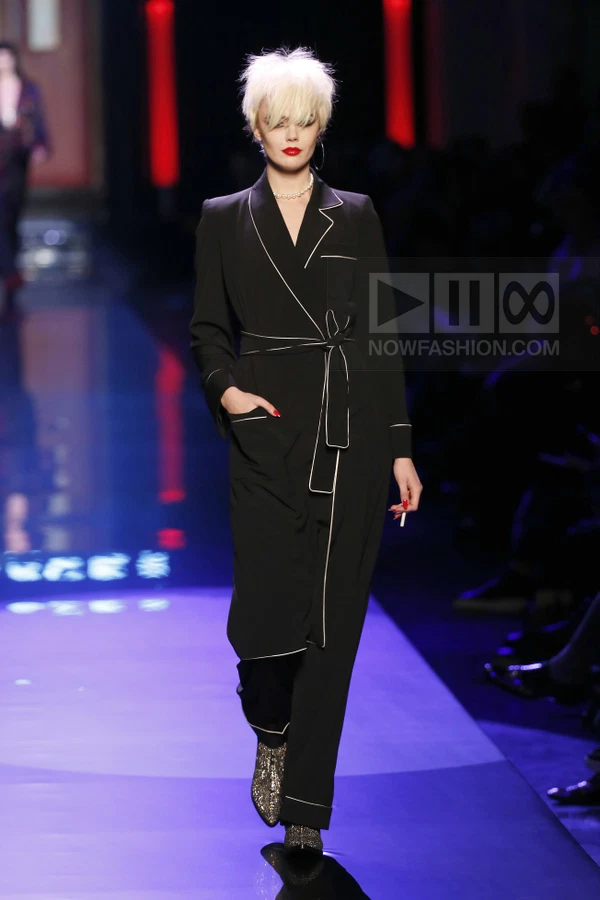 Model walking in a black wrap coat with white contrast piping, worn over black trousers and metallic boots on a runway.