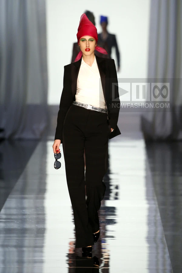 Model walking on a runway wearing a black tuxedo jacket, white shirt, black trousers, and a bright pink headscarf.
