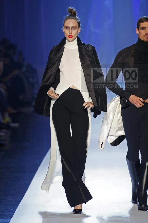 Model wearing a white turtleneck top, black trousers, and a black jacket draped over shoulders on a runway.