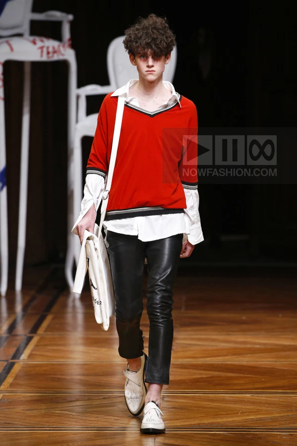 A person walks the runway wearing a red v-neck sweater over a white shirt, black leather pants, and white shoes.