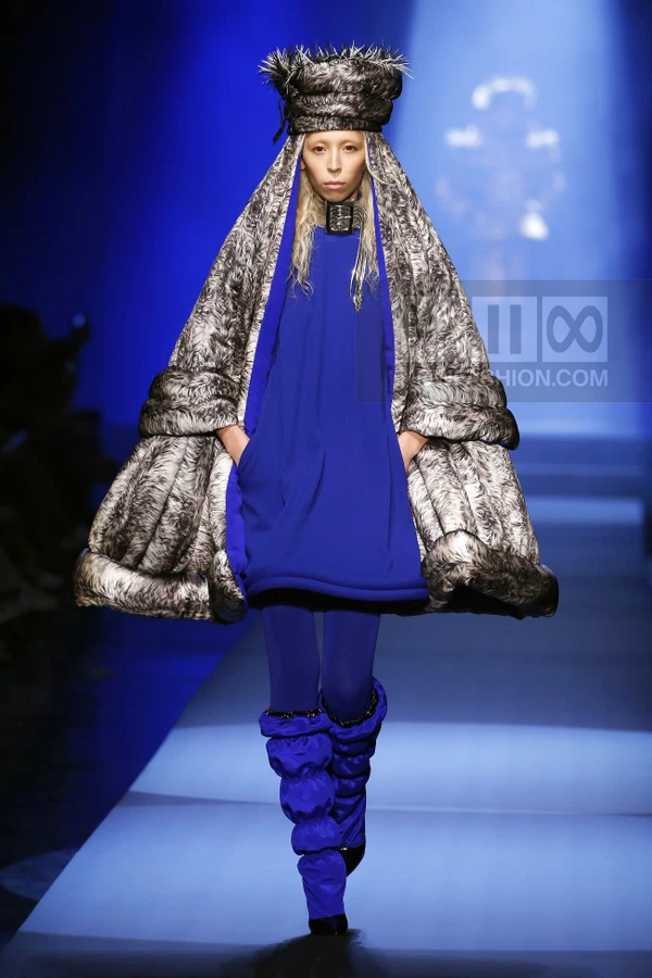Model on runway wearing a blue mini dress, oversized faux fur cape, matching blue leggings, and a tall fur hat.