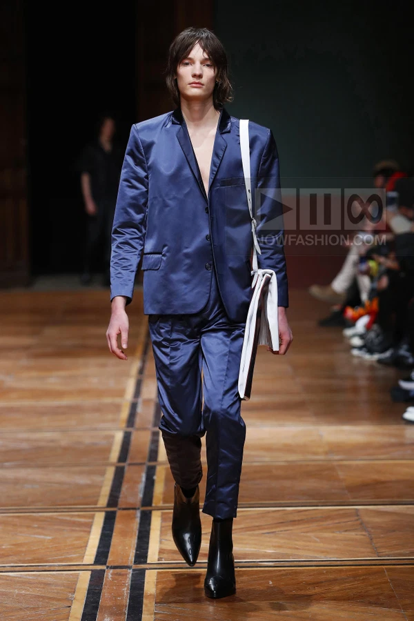 A person walks a runway wearing a navy blue satin suit with a white shoulder bag and black pointed-toe boots.