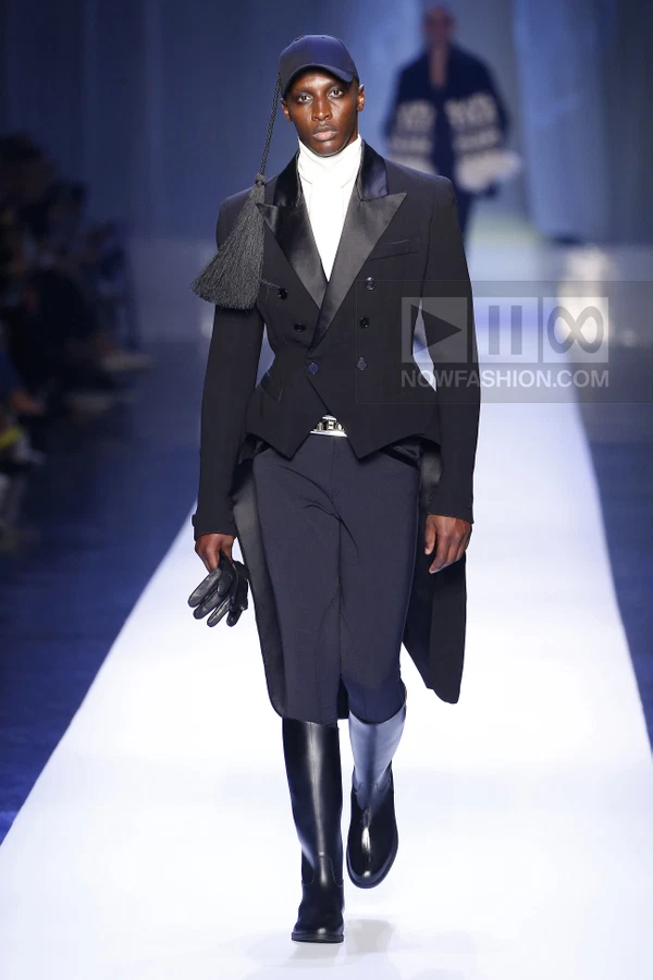 Model wearing a black tuxedo tailcoat, white turtleneck, and baseball cap with a long tassel on a runway.