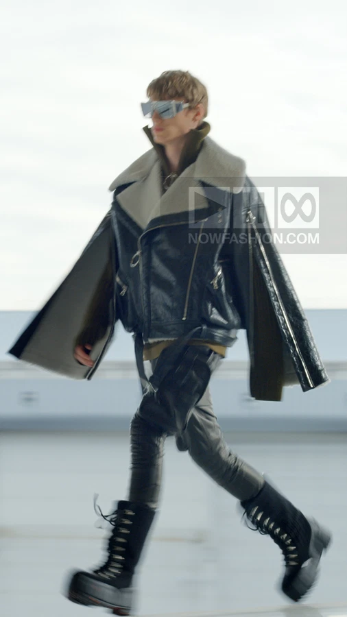 Model walking in a black leather biker jacket with shearling collar, olive sweater, leather pants, and black combat boots.