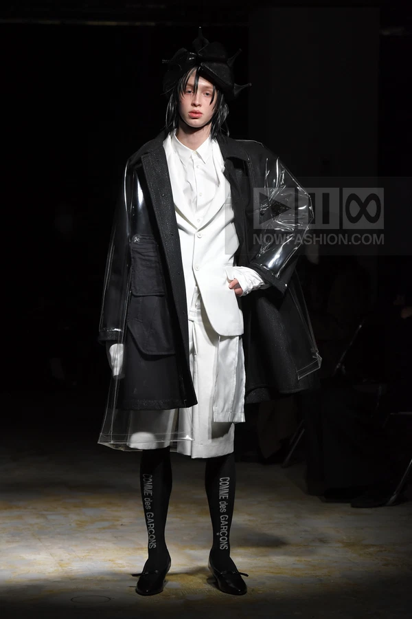 Model wearing a white suit, black coat with clear vinyl overlay, black headwear, and branded black tights on a runway.