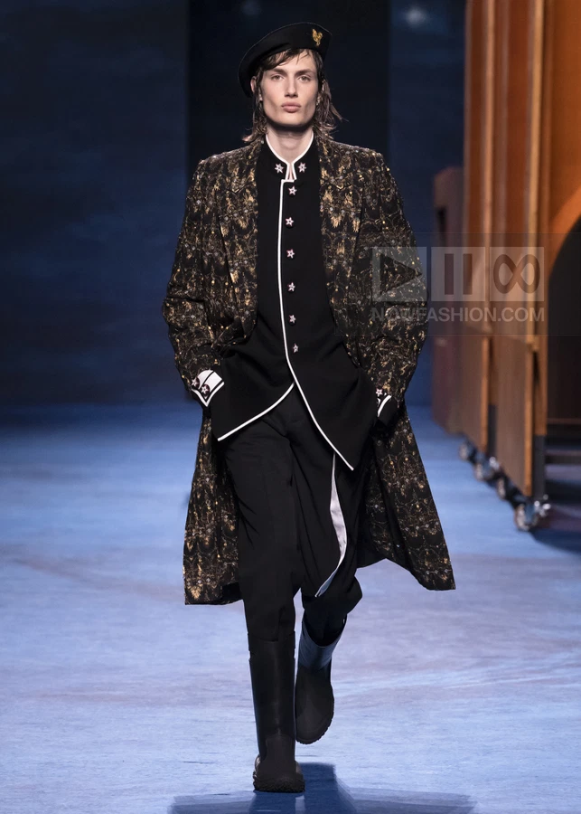 Model walking in a black and gold patterned coat, black jacket with white trim, black trousers, and black boots.