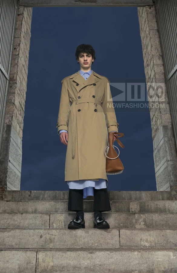 A person stands on stone steps wearing a beige trench coat over a light blue shirt and black cropped trousers.