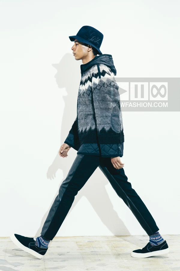 A person walking in a patterned knit hooded jacket, dark trousers, bucket hat, and slip-on sneakers against a white wall.