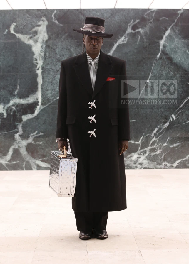 A person wearing a black overcoat with airplane-shaped buttons, a fedora, and carrying a metallic silver briefcase.
