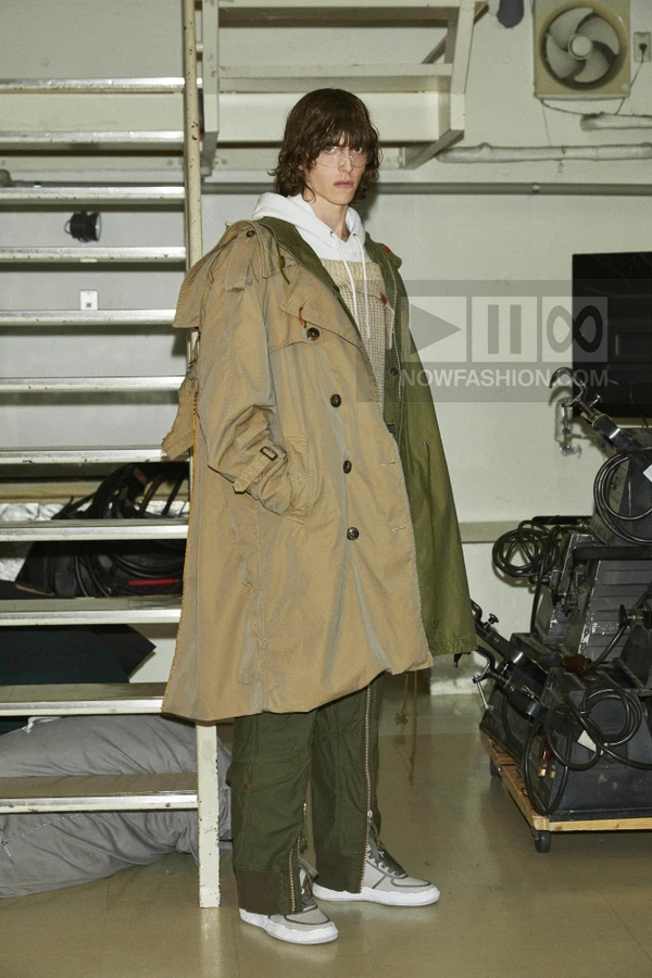 Person wearing a layered khaki and olive green coat over a white hoodie, textured sweater, and olive cargo pants.